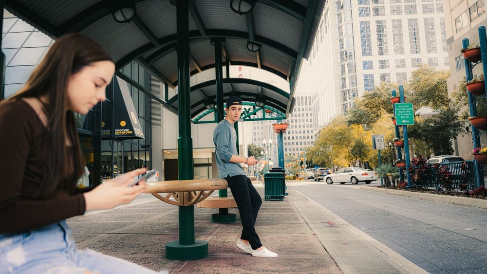 Two teenagers at a city bus shelter, sitting apart, each looking down at their own phone