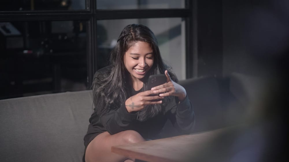 Una giovane donna seduta su un divano, in una stanza in penombra, sorride guardando il telefono