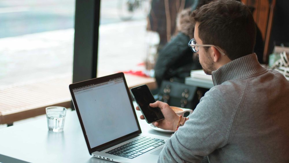 Un homme consulte son téléphone devant son ordinateur portable ouvert, à une table près d'une fenêtre