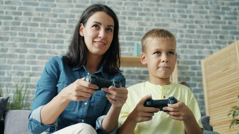 A mother and her son, controllers in hand, playing a video game together on the living room sofa