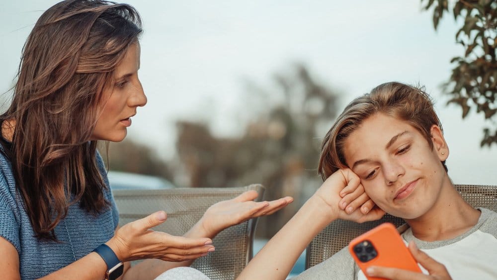 Una madre parla con il figlio adolescente seduto accanto a lei al tavolino di un bar, mentre lui tiene gli occhi bassi sul telefono