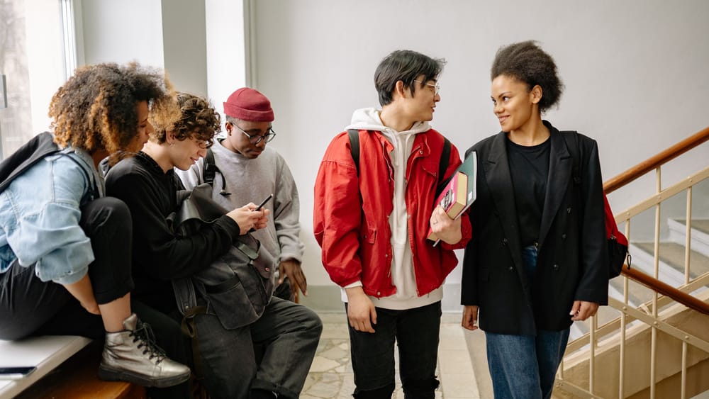 Students on a staircase landing: one group around a phone, two others talking, books in hand