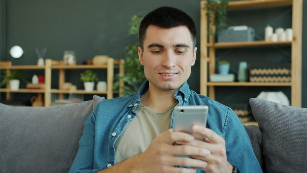 Un homme souriant assis sur un canapé, en train de regarder son téléphone